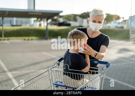 Mutter hilft ihrem Sohn, der in einem Einkaufswagen sitzt, eine Schutzmaske zu tragen, bevor er in einen Laden kommt. Stockfoto