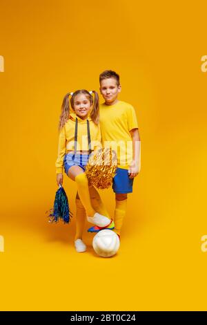 Ein Cheerleader und ein Fußballspieler in der gleichen Uniform stehen und halten den Ball mit dem Fuß. Mädchen mit Pompons Stockfoto