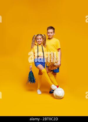 Ein Cheerleader und ein Fußballspieler in der gleichen Uniform stehen und halten den Ball mit dem Fuß. Mädchen mit Pompons Stockfoto