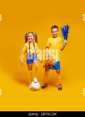 Nettes Cheerleader-Mädchen und ein Fußballspieler in der gleichen Uniform stehen, halten den Ball mit dem Fuß. Junge, die Pompons halten Stockfoto