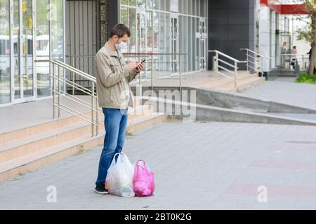 Ein Mann in einer medizinischen Maske und ein Einkauf hielten auf einer leeren Straße an, um einen Anruf anzunehmen Stockfoto