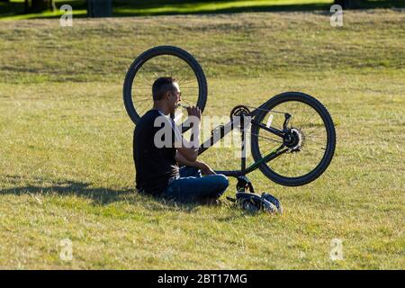 Ein Mann mit Radler entspannt sich in einem öffentlichen Park auf dem Gras und genießt den sonnigen Sonnentag mit seinem Fahrrad auf den Kopf gestellt. Er raucht ein nicht-Tabakgerät, wie eine Vaping-Zigarette London Middlesex UK (119) Stockfoto