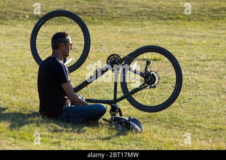 Ein Mann mit Radler entspannt sich in einem öffentlichen Park auf dem Gras und genießt den sonnigen Sonnentag mit seinem Fahrrad auf den Kopf gestellt. London Middlesex Großbritannien (119) Stockfoto