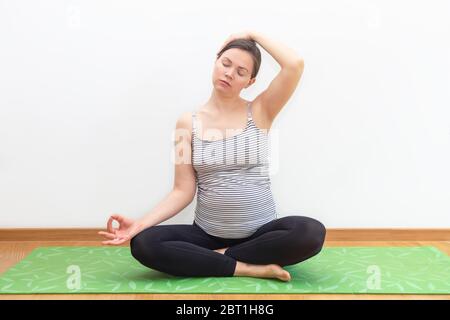 Pränatales Yoga- und Fitnesskonzept. Schwangere Frau in halb Lotushaltung Training zu Hause Stockfoto