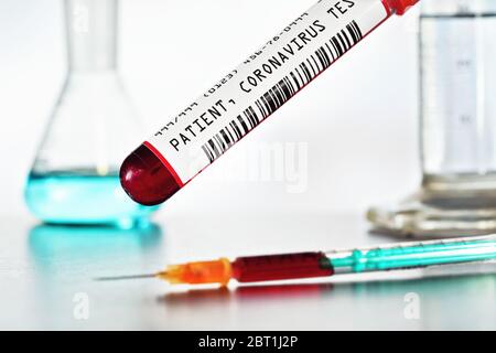 Probenflasche mit Blut, Etikett (eigenes Design, Dummy-Barcode und Daten) Coronavirus-Test, grüne orangene Spritze oben. Hintergrund der Laborausrüstung verwackelt Stockfoto