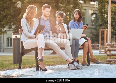 Besorgt kaukasischen Geschäftsleute sitzen im Park auf Bank, arbeiten auf Laptop, Handys, Tablet, während ihrer Pause. Geschäft nach Corona-Virus, Stockfoto