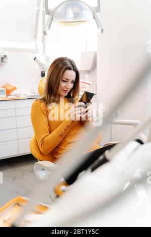 Junge Frau lächelt und mit dem Smartphone, während sie auf dem Stuhl sitzt und auf den Arzt in der modernen Zahnarztpraxis wartet Stockfoto