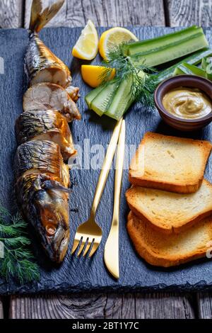 Hausgemachte, geräucherte, heiße Makrele auf einem schwarzen Schneidebrett mit dijon-Senf, frischen Gurken, Dill, Toasts, Zitronenpfeilchen und Besteck auf einem Holzbrett serviert Stockfoto