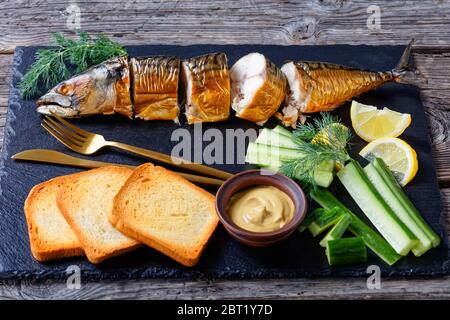 Fettiger Fisch: Heiße geräucherte Makrele auf einem schwarzen Schneidebrett mit dijon-Senf, frischer Gurke, Toasts, Zitronenpfeilchen und Besteck auf einem Holzbackgr serviert Stockfoto