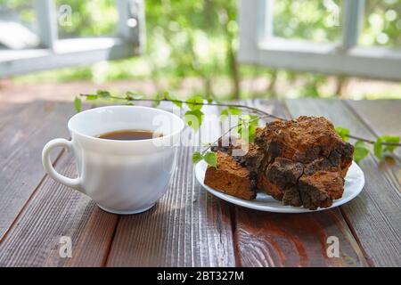 Chaga Pilz und eine Tasse Tee stehen auf einer Holzoberfläche neben einem offenen Fenster an einem sonnigen Sommertag. Stockfoto