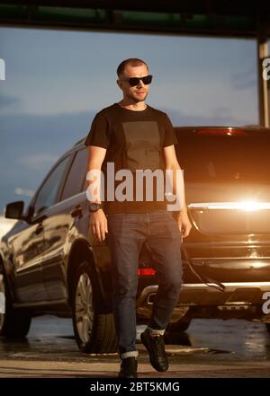 Portrait eines erfolgreichen Mannes mit Sonnenbrille, schwarzem T-Shirt, dunkelblauer Jeans und schwarzen Sneakers läuft neben seinem schwarzen Auto, in voller Länge Stockfoto