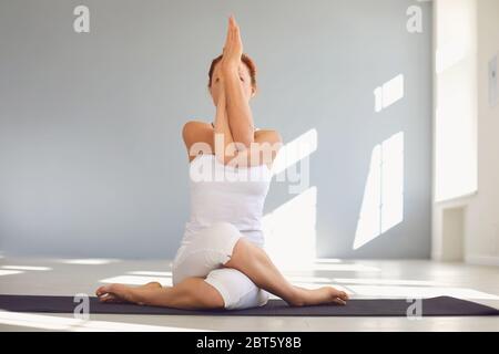 Junge fokussierte Frau in weißer Sportbekleidung, die sitzende Eagle-Übung oder Garudasana-Pose durchführt, während sie Yoga im hellen Raum praktiziert Stockfoto