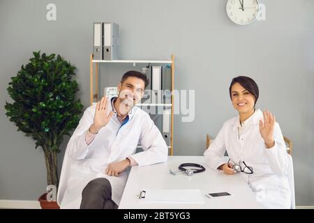 Freundliche Ärzte, die am Tisch sitzen und in einer modernen Klinik arbeiten, lächeln und die Hände winken Stockfoto