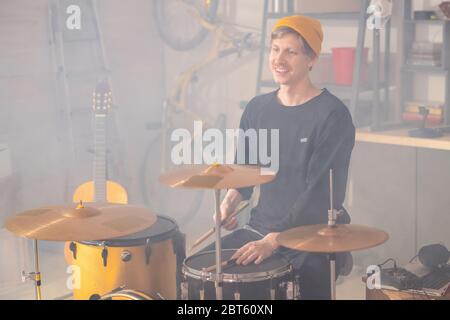 Fröhlicher junger, schöner Musiker in gelber Beanie und schwarzer Casualwear, der während der Probe in der Garage oder im Studio vor dem Schlagzeug sitzt Stockfoto