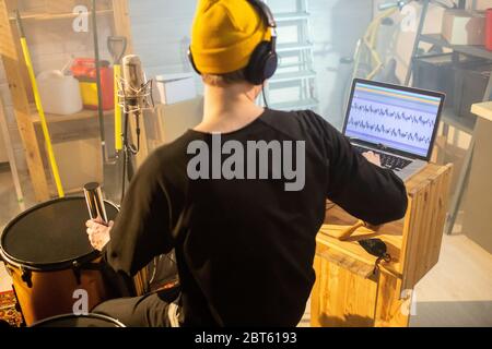 Rückansicht des jungen Musikers in Kopfhörern, der Musik vor Laptop, Mikrofon und Schlagzeug aufzeichnet, während er in der Garage oder im Studio sitzt Stockfoto