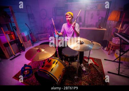 Junger Mann in Freizeitkleidung, der die Hände mit Schlagstöcken hebt, während er während der Probe auf Becken trifft und Musik in der Garage oder im Studio aufnimmt Stockfoto