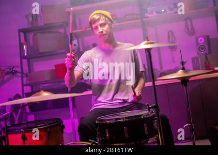 Typ in T-Shirt, Jeans und Mütze sitzt in der Garage vor dem Schlagzeug und geht auf Becken und Trommel mit Drumsticks während der Probe zu schlagen Stockfoto