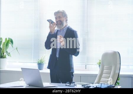Glücklicher älterer Mann im eleganten Anzug, der Sprachnachrichten mit dem Smartphone am Mund aufzeichnet, während er am Arbeitsplatz im Büro steht Stockfoto