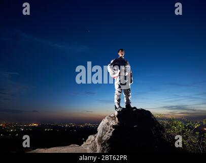 Tapferer Astronaut auf dem felsigen Hügel mit schönen blauen Himmel auf dem Hintergrund. Schöner Spaceman in weißen Raum Anzug hält Helm. Konzept der Kosmonauten und Raumfahrt. Stockfoto