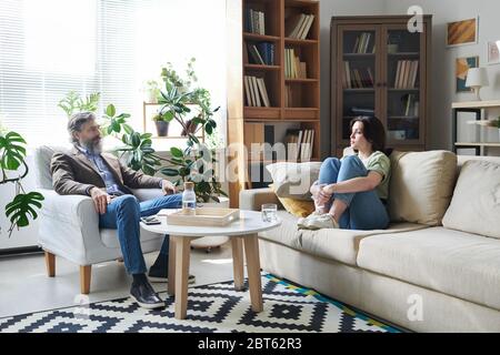 Junge unruhige Frau in Freizeitkleidung sitzt auf der Couch im Büro des Beraters sitzt vor ihr und spricht mit ihm über ihre Probleme Stockfoto