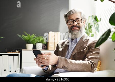 Reife grauhaarige erfolgreiche Profi in Brillen und formalwear Sitzen in weißen Ledersessel und Scrollen in Handy Stockfoto