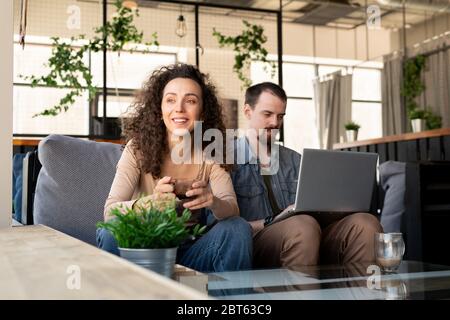 Glückliche junge erholsame Frau mit einer Tasse Tee oder Kaffee, die durch das Fenster schaut, während sie auf der Couch neben ihrem Mann mit Laptop sitzt Stockfoto