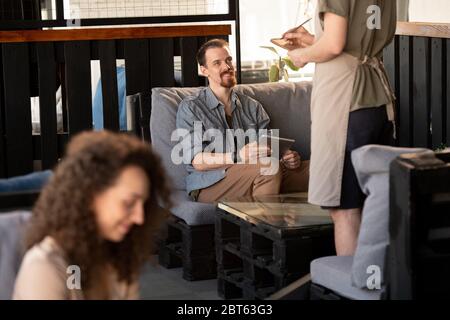 Fröhlicher junger bärtiger Mann in Freizeitkleidung, der Ordnung im Restaurant oder Café macht, während er auf die Kellnerin schaut, die vor ihm steht Stockfoto