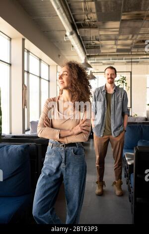 Fröhliche junge Frau in beigem Pullover und blauen Jeans, die Arme an Brust kreuzt, während sie am Sofa steht und durch das Fenster im Café schaut Stockfoto