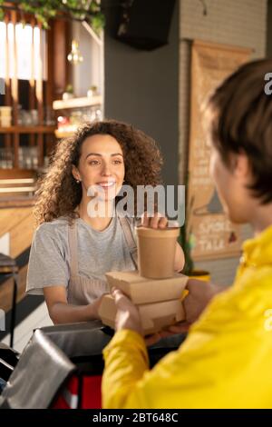 Junge lächelnde Kellnerin, die auf Kurier schaut, der vor ihr steht, während sie ihm hilft, Aufträge von Kunden in große Tasche im Restaurant zu packen Stockfoto
