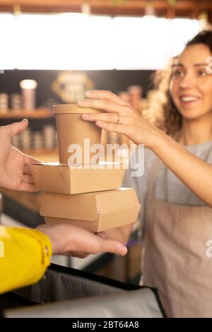 Hand der jungen Kellnerin in Schürze hält zwei Karton-Behälter mit Lebensmitteln und Glas Kaffee über offene Tasche, während sie an Kurier übergeben Stockfoto