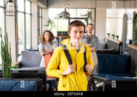 Junger Kurier in gelber Jacke, der Rucksack hält, während er im Gang vor der Kamera steht, mit zwei fröhlichen Kellnern im Hintergrund Stockfoto
