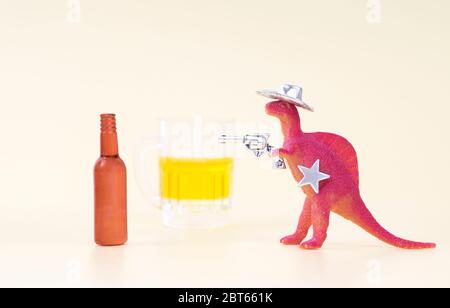 Spielzeug-Dinosaurier mit Cowboy-Hut, hält einen Revolver in seinem Arm und mit einem Sheriff-Stern schießt eine leere Bierflasche auf einem neutralen Hintergrund. Kämpfen Stockfoto