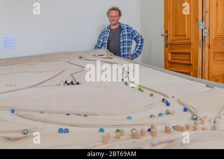 23. Mai 2020, Brandenburg, Altranft: Reinier Scheers, Tischler und Designer des Modells, steht an seiner Marmorbahn im Oderbruch Museum Altranft. Reinier Scheers Aufgabe war es, das Strömungssystem des Oderbruchs mit Glasmurmeln sinnlich erlebbar zu machen. Das Ergebnis ist eine einzigartige Marmorbahn einer Region in Ostbrandenburg. Die Marmorbahn wird nicht mit elektrischer Energie betrieben. Nur Mechanik und Schwerkraft treiben dieses neue Highlight des Museums an. Und wer das Museum kennt, weiß, dass hier alles berührt werden kann oder sollte: Lernen durch Spielen, Lernen durch Berührung ist das Programm dort. Th Stockfoto