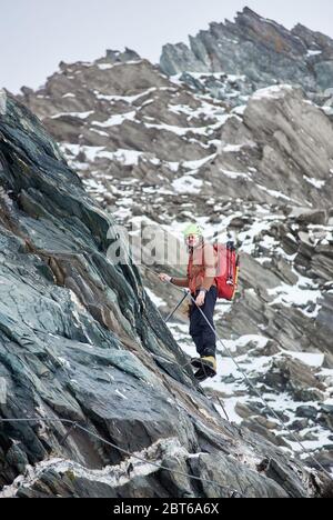 Männlicher Alpinist in Sonnenbrille und Schutzhelm mit Rucksack aufsteigenden hohen Berg. Mann Kletterer mit festem Seil und Blick auf die Kamera. Konzept des Bergsteigens, Alpinismus und Kletterns. Stockfoto