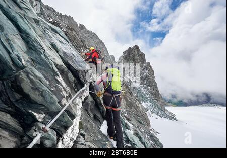 Rückansicht von Bergsteigern mit Rucksäcken mit Fixseil beim Aufstieg hohen felsigen Berg. Männliche Bergsteiger klettern natürliche Felsformation und versuchen, Berggipfel zu erreichen. Stockfoto