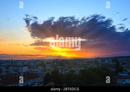 Die Strahlen der untergehenden Sonne brechen durch eine große Wolke, während die Sonne über dem Horizont untergeht. Goldener blauer Abendhimmel über einer kleinen Stadt. Sommer Skysca Stockfoto