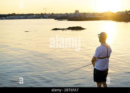 Fischer, die am Meer stehen, Haken einen Fisch gegen Sonnenuntergang. Sportler hält eine Angelrute und Haspel in einem Haken Fisch. Spin Angeln Hobby Fang ein Stockfoto