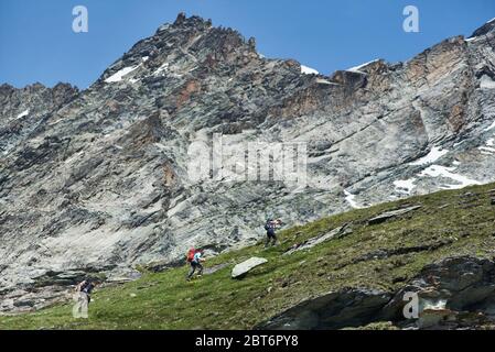 Seitenansicht der Gruppe von drei Touristen mit Trekking-Ausrüstung zu Fuß auf dem Weg, schöne Berglandschaft im Hintergrund. Bergwandern, Männer erreichen den Gipfel im Sommer Tag. Sporttourismus in den Alpen. Stockfoto