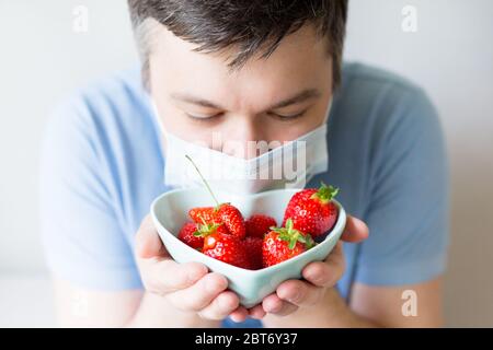 Junger Mann in medizinischen Maske hält Schüssel mit Erdbeere. Mann in einer Anti-Virus-medizinischen Maske. Erholung von Coronavirus. Stoppen Pandemie Stockfoto