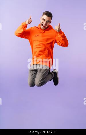 Foto von positiven emotionalen jungen Mann springen isoliert über lila Hintergrund trägt Brillen mit Daumen nach oben Geste. Stockfoto