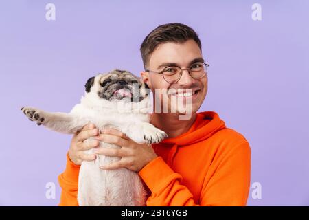 Bild von glücklichen positiven Mann posiert isoliert über lila Hintergrund halten umarmenden Hund Mops. Stockfoto