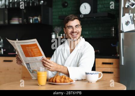 Lächelnder junger Mann, der bademantel trägt und morgens in der Küche sitzt und die Zeitung liest Stockfoto