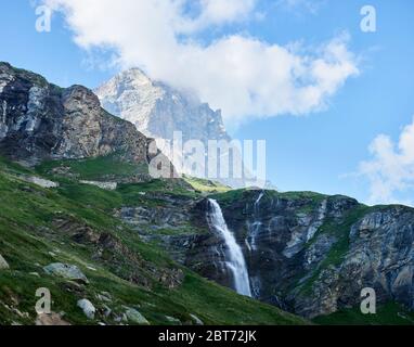 Landschaft von Wasserfall in Bergtal unter blauem bewölktem Himmel. Malerisches Panorama von grasbewachsenen Hügeln, Wasserfall, Berggipfel Matterhorn umgeben von weißen Wolken. Konzept der Schönheit der Natur. Stockfoto