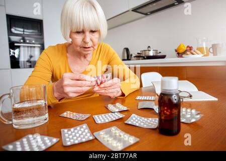 Ruhestand. Ältere Frau sitzt am Tisch in der Küche unter Blister mit Pillen Gefühl unwohl Nahaufnahme besorgt Stockfoto