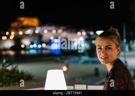 Blonde Frau dreht das Gesicht und schaut auf die Kamera, während sie auf einer Terrasse lächelt bei Nacht mit den Lichter der Stadt außerhalb des Fokus im Hintergrund Stockfoto