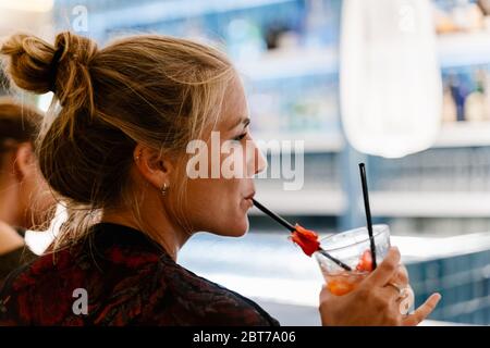 Profil einer blonden Frau auf dem Rücken, die an einer Bar sitzt und einen Strohhalm eines Cocktails lutscht Stockfoto