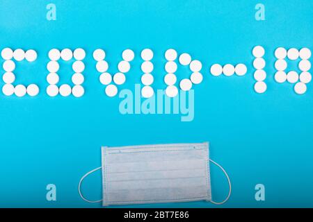 Die Inschrift COVID-19 aus weißen medizinischen Pillen und schützender medizinischer Maske liegt auf einem cyanfarbenen Papierhintergrund. Konzept zur Impfstoffentwicklung. Große Pharmaindustrie. Stockfoto