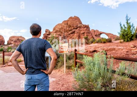 Canyons im Arches National Park mit Skyline Arch im Hintergrund und dem Rücken eines jungen Mannes, der auf der Trailhead Trail Wanderung in Utah steht Stockfoto