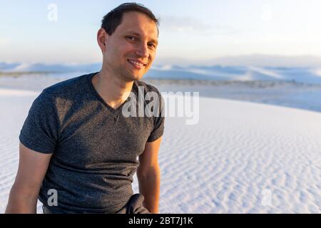 Porträt eines jungen Mannes, der auf Sand am White Sands Dunes National Monument, New Mexico sitzt und die Kamera während des Sonnenuntergangs über dem Horizont mit dem Orgelberg betrachtet Stockfoto