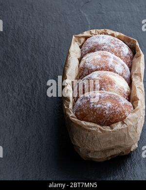 Frisch gebackene Donuts gefüllt mit Erdbeermarmelade in Papiertüte auf schwarzem Steingrund Stockfoto